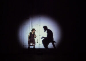 Nervous Theatre's Isabel Shaida sits on a chair on stage with the shadow of a bearded man beside her.