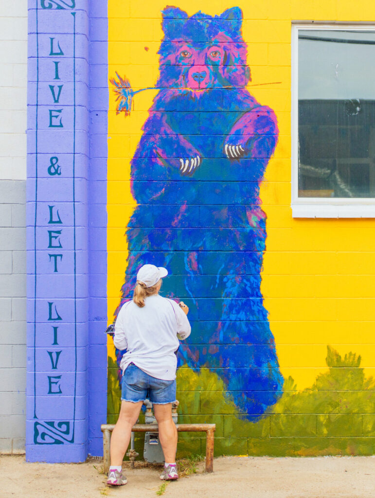 Artist Mary Arnold works on her mural, Live and Let Live, which features a brightly colored blue bear on a yellow background.